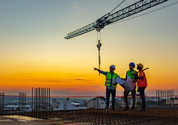 Three people stand on top of a building wearing safety gear, with a crane and an orange sky in the background. 