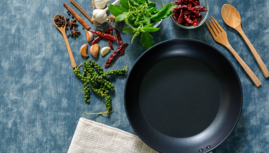 Nonstick frying pan surrounded by fresh herbs, spices, garlic, and wooden kitchen utensils on a blue surface.