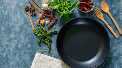 Nonstick frying pan surrounded by fresh herbs, spices, garlic, and wooden kitchen utensils on a blue surface.